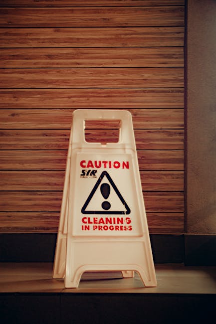 A white plastic 'Caution Cleaning in Progress' floor standing sign with a black triangle warning symbol and red text, positioned on a grey tiled floor against a background of wooden wall paneling and