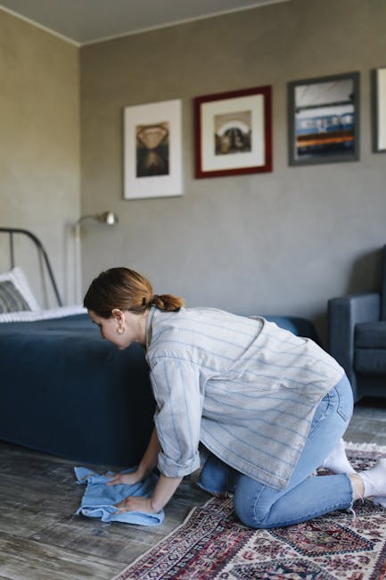 A woman, with curly hair tied back and wearing a beige jacket and blue jeans, is engaged in typical domestic surface cleaning using a yellow vacuum cleaner with a black hose. She is standing on a patt