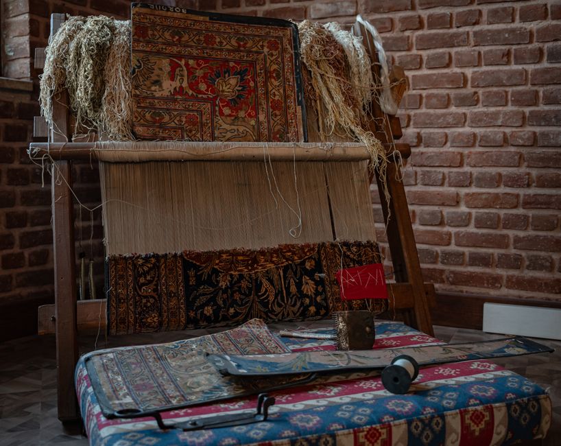 A display of various antique and traditional rugs and carpets arranged outdoors on a wooden platform and the ground, showcasing intricate patterns, vibrant colors, and detailed craftsmanship. Some rug